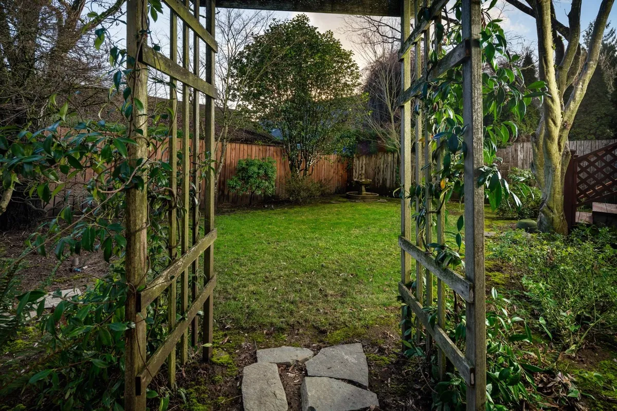 A wooden arbor is surrounded by lush greenery, with a stone pathway meandering through a peaceful garden. The scene conveys a sense of tranquility and privacy, suggesting a quiet retreat just steps from home.