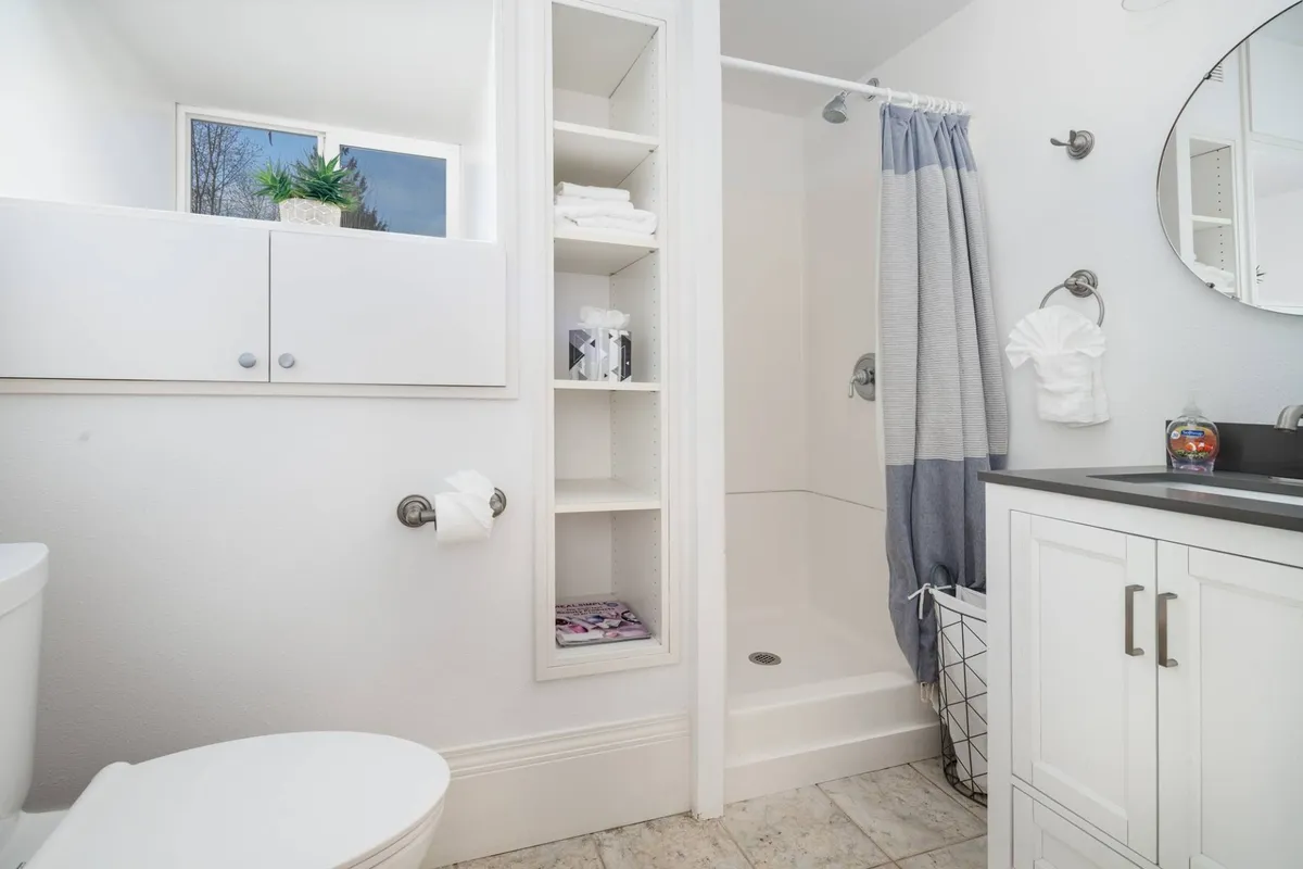 A bright, modern adjacent bathroom featuring a toilet, shower, sink, and storage shelf, designed for comfort and convenience during gatherings or family meals.