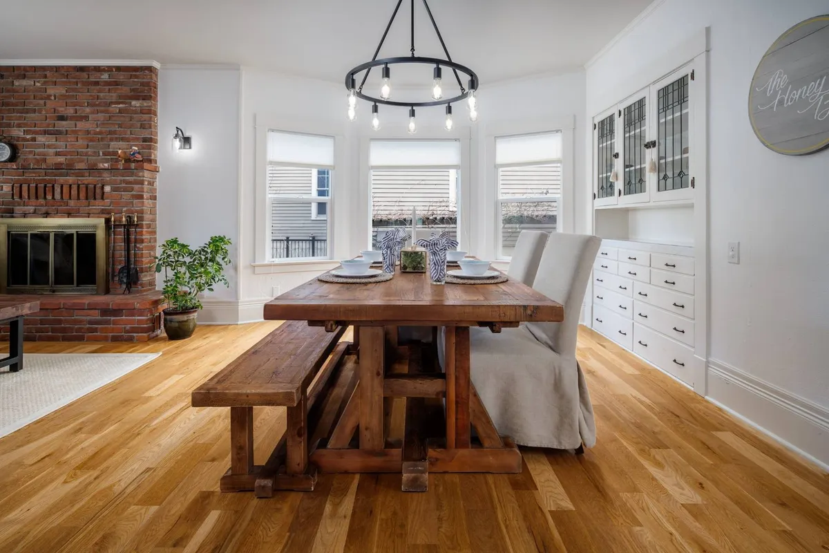 This image shows a warm, inviting dining space with a wooden table set beneath a chandelier. Large windows bring in natural light, and a brick fireplace adds to the homey atmosphere. The setup suggests a welcoming environment ideal for family meals and gatherings.