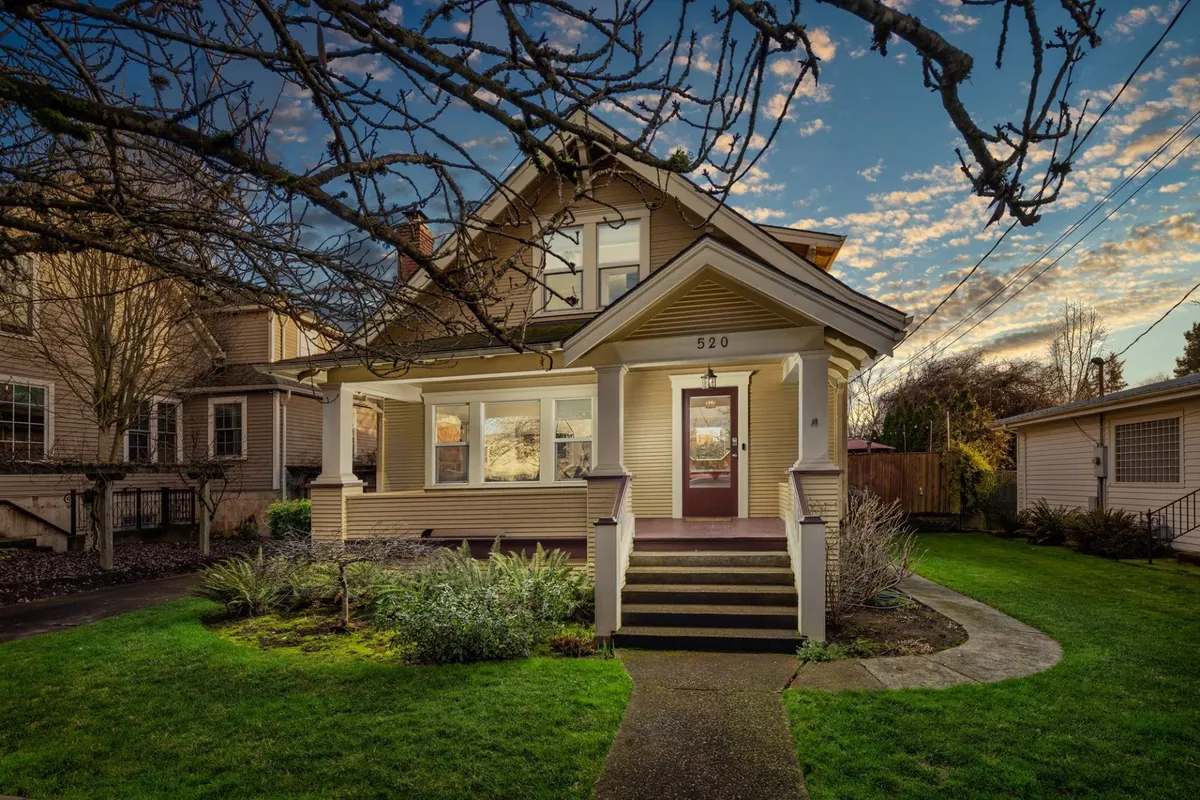 The image showcases the inviting front porch and entrance of a classic 1920's craftsman home, framed by lush gardens and bathed in the warm glow of sunset. Comfortable seating hints at gatherings with family or neighbors, while the well-kept yard is perfect for outdoor relaxation or a sunset barbecue.