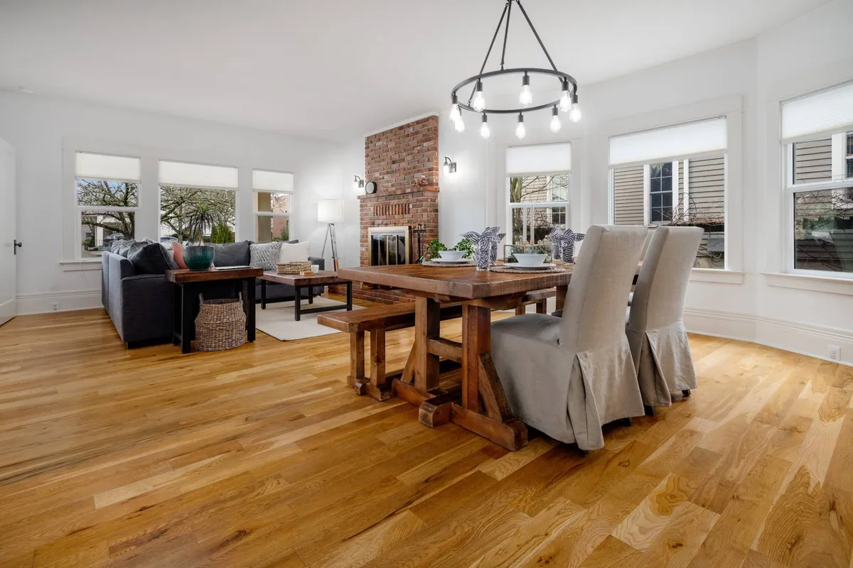 This image features a spacious and welcoming living area with a prominent dining table, a cozy sofa set near a glowing fireplace, and a stylish chandelier overhead. The space is arranged to encourage group gatherings, with plenty of room for guests to mingle, dine, or relax together.