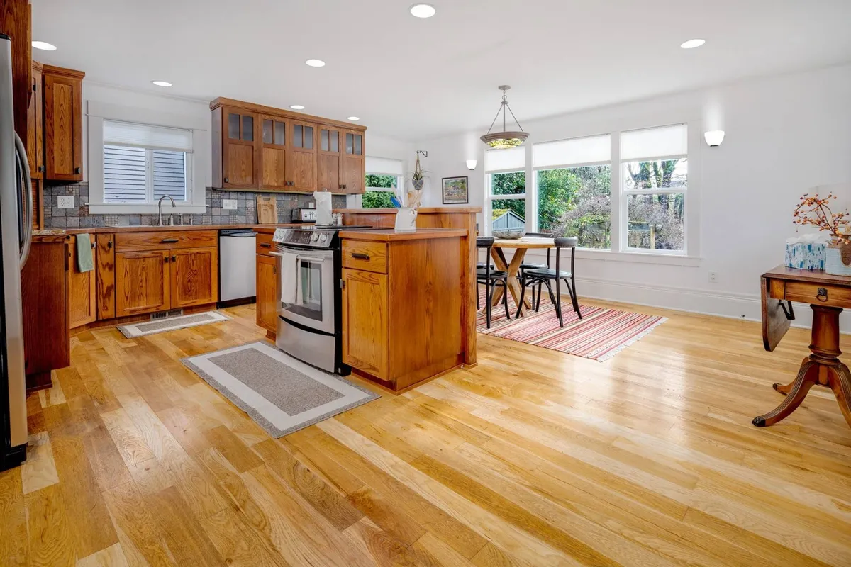 This image showcases a spacious kitchen featuring wooden cabinets, a central kitchen island, a dining table, and large windows that flood the space with natural light. The layout suggests both functionality and sociability, ideal for cooking and gathering with family or friends.