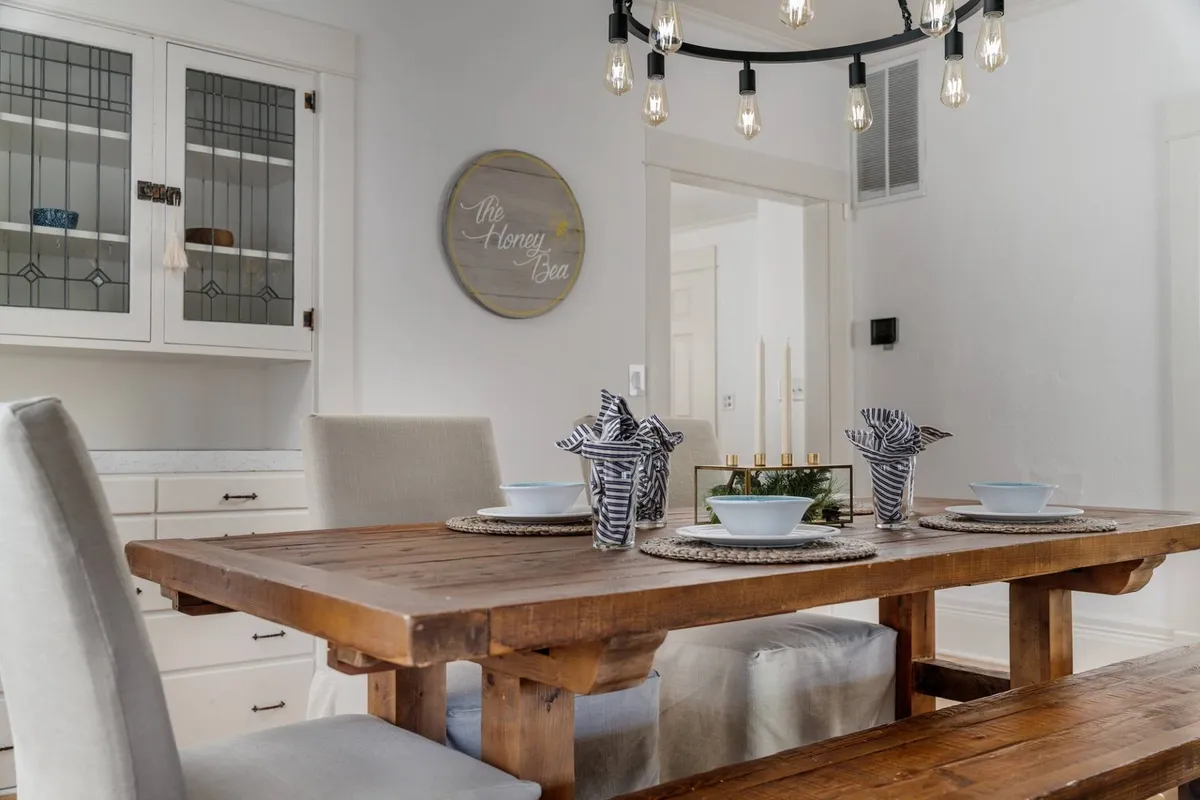 The scene spotlights a large wooden dining table under an eye-catching chandelier with exposed bulbs, accompanied by a decorative wall sign. The table is set for a meal, epitomizing the warmth of shared dining experiences and lively conversations among friends or family.