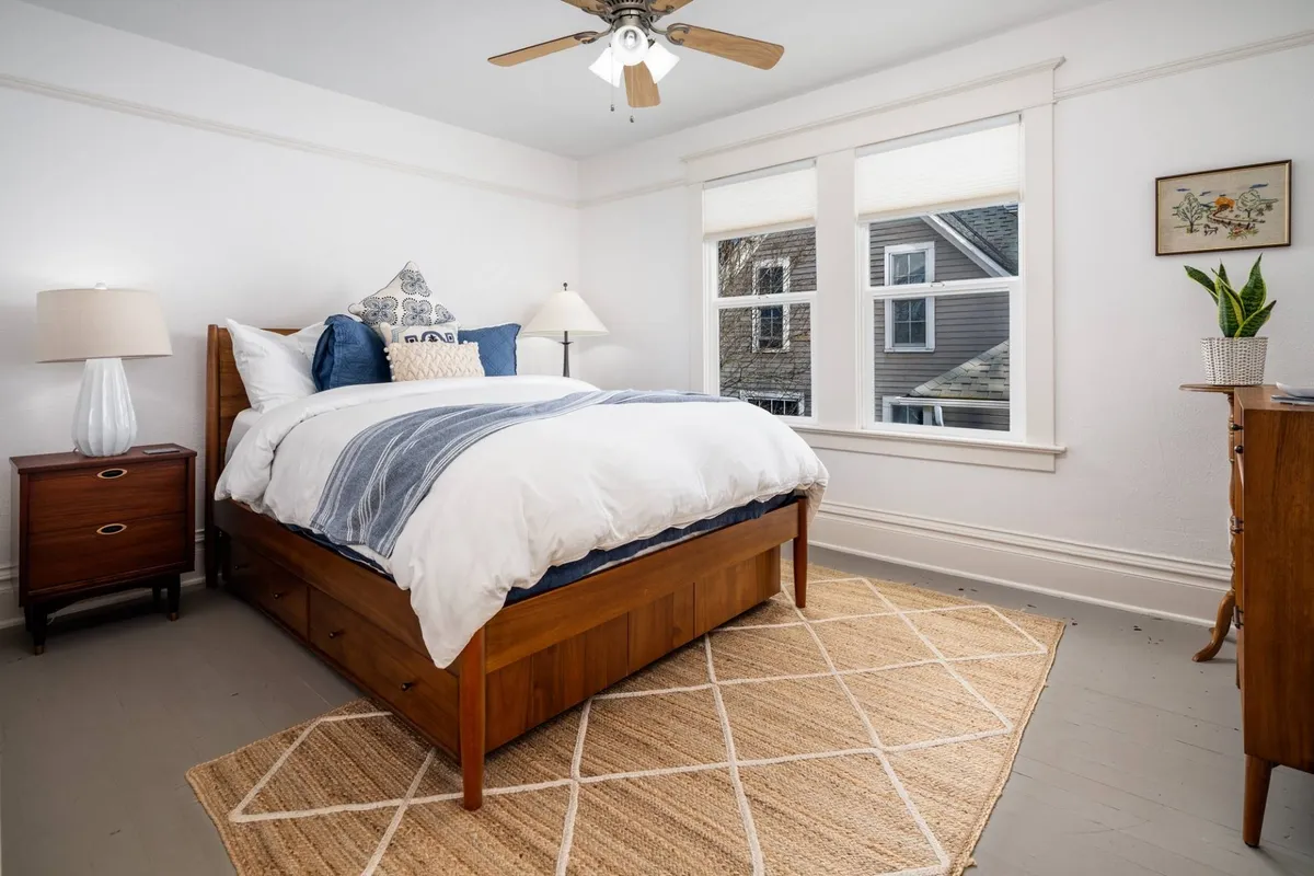 A serene bedroom with a queen bed, soft lighting from bedside lamps, plush bedding, and natural light pouring in through windows. A dresser adds practical storage, and a rug brings warmth to the space.