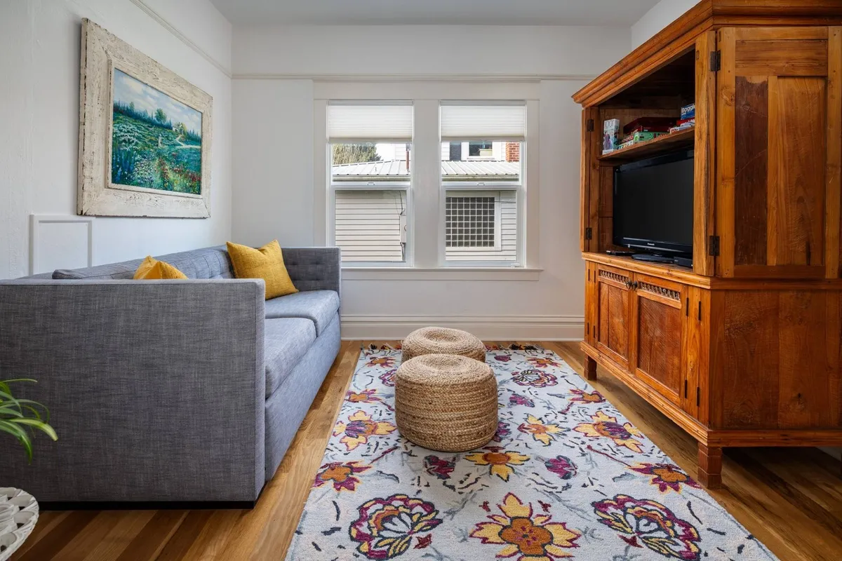A comfortable gray sofa faces a wooden cabinet with a colorful area rug underfoot and artwork on the wall. The room radiates a cozy and welcoming vibe, suitable for lounging, socializing, or hosting movie nights with friends and family.